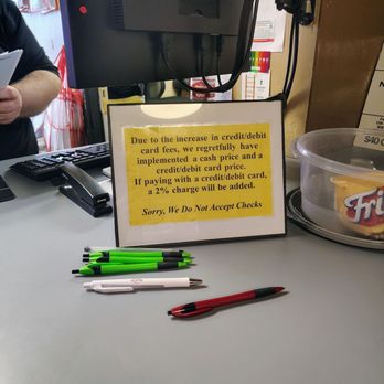 Network Automotive Service Center - Enlarge to see the surcharge on debit cards. BREAKS FEDERAL LAWS - A yellow printed sign on a business counter indicates that due to increased credit and debit card fees, a cash price and a credit/debit card price are implemented, with a 2% surcharge for card payments. The sign also states that checks are not accepted. The counter has several pens, a computer monitor, and a plastic container with a snack inside. The background includes part of a person and some office or store equipment. - Auto Repair Near Me - Apache Junction, Arizona