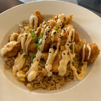 Le Saigon - Boom Boom Chicken over Fried Rice - A bowl contains fried chicken pieces served over a bed of rice. The dish is topped with mayonnaise drizzles, black sesame seeds, and chopped green onions, presented in a white bowl. The setting appears to be a casual dining environment, emphasizing Asian-inspired cuisine. - Asian Fusion Near Me - Hammond, Louisiana