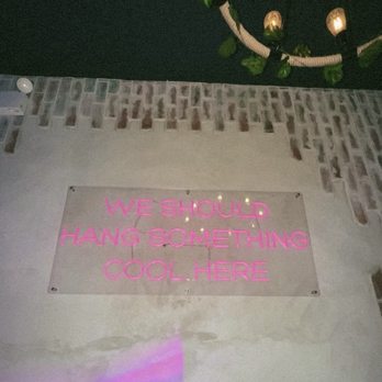 Theme House - A neon sign with pink lettering is mounted on a wall, displaying the message 'We should hang something cool here.' The wall has a brick pattern at the top and a plain surface below. - Karaoke Near Me - Chicago, Illinois