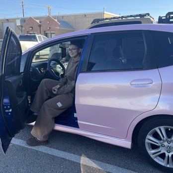 The Wrap Lab - 2010 Honda Fit, revitalized by the Wrap Lab.  Color Sakura pink. Thank you Wrap Lab for a great application of the car wrap. - A woman wearing glasses and a brown outfit sits in the driver's seat of a pink Honda Fit in a parking lot. - Vehicle Wraps Near Me - Albuquerque, New Mexico