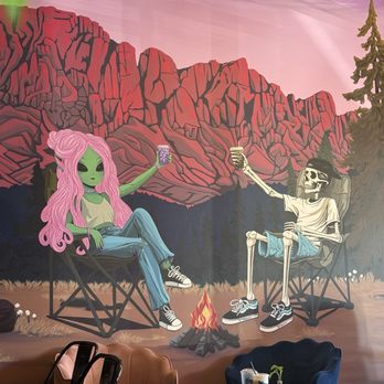 Wild Pine - The mural features an alien girl with green skin and long pink hair, sitting on a camping chair, holding a jar. Next to her, a skeleton wearing a hat and sunglasses sits on a similar chair, both extending their arms towards each other with jars in hand. A small campfire is in the foreground, with a mountain range and starry sky painted behind them. The scene is set outdoors with trees on the right side, creating a camping atmosphere. - Coffee & Tea Near Me - Peoria, Arizona