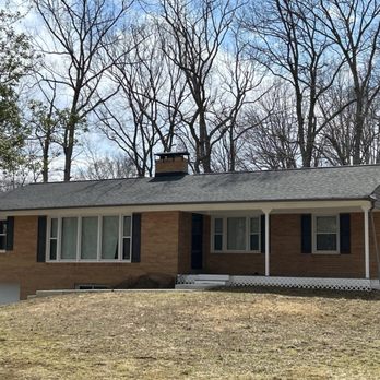 RoofPRO - A single-story brick house features a sloped roof, several windows with black shutters, and a small front porch with steps leading up to the entrance. The house is set on a grassy yard with a slight incline, and leafless trees are visible in the background, indicating a winter or early spring season. - Roofing Near Me - Severn, Maryland