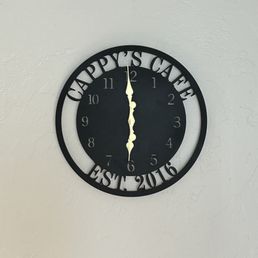 Nice Clock, set 2016 and still going strong. Everyone gives her clocks for some reason. Take her one. - A black, round wall clock with cut-out text 'HAPPY'S CAFE' at the top and 'EST. 2016' at the bottom, mounted on a plain, light-colored wall.