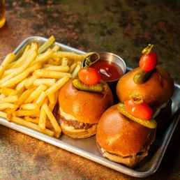 Wagyu Slider W/ Foie Gras - Three sliders with buns, cherry tomatoes, and pickles are arranged on a metal tray. Each slider contains a beef patty and cheese, topped with a cherry tomato and a pickle. A small metal container of ketchup is placed nearby. The tray is set on a textured, dark-colored surface, with two glasses partially visible in the background.