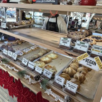 Le Croissant - A bakery display case contains various baked items such as cookies, croissants, muffins, and danishes, with labels indicating flavors and vegan options. - Bakeries Near Me - Logan, Utah