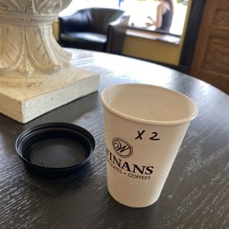 Cup o Joe - A white paper coffee cup with the Winans logo and 'x 2' written on it is positioned on a dark wooden table inside a cafe. The cup is accompanied by a black plastic lid placed upside down. In the background, a large decorative white vase is visible on the table, and through the window, a woman is seen sitting and using her phone, with outdoor scenery and sunlight illuminating the scene.