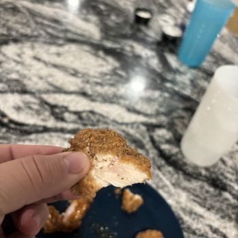 Buffalo Wild Wings - RAW CHICKEN - A hand is holding a piece of fried chicken with a golden-brown, crispy coating, revealing the white meat inside. Several other pieces of fried chicken are on a dark blue plate beneath the hand. The background shows a marble countertop with a blue cup, a white cup, and some small containers, indicating a kitchen or dining area setting. - Chicken Wings Near Me - Conroe, Texas