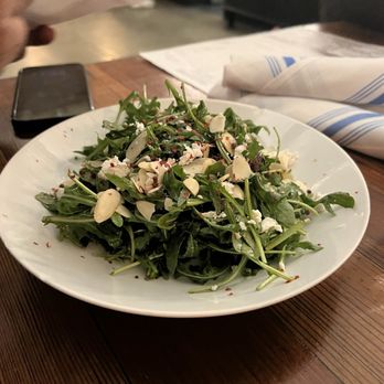 Forbidden Root - Arugula Salad - A white, round plate holds a fresh arugula salad garnished with sliced almonds and crumbled cheese. The salad appears vibrant and crisp, with the ingredients evenly distributed. The plate is placed on a wooden table, with a smartphone, napkin, and some papers visible nearby. The setting suggests a casual dining environment, possibly in a restaurant or cafe. - Brewpubs Near Me - Chicago, Illinois