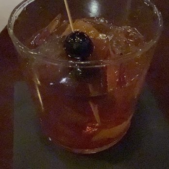 Steak + Vine - Cocktail - A cocktail with ice cubes and a dark cherry garnish is presented in a clear glass. The drink appears to be a dark-colored beverage, and the glass is set on a dark surface, likely a table or bar counter. The background is dimly lit, suggesting an indoor or nighttime setting. - Steakhouses Near Me - La Grange, Illinois