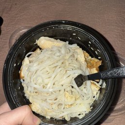 A black plastic takeout bowl holds cooked flat noodles, scrambled eggs, and bread pieces, with a black spoon inside, on a brown textured surface.