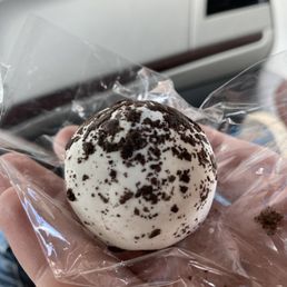 Oreo Cake Pop - A cookie with white icing and chocolate crumbs is held in a person's hand, wrapped in clear plastic, with a vehicle interior in the background.