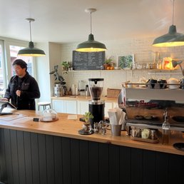 Faro cafe interior featuring a wooden counter, pendant lights, a menu board, and a barista preparing an order.