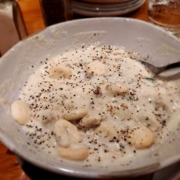 Clam Chowder - A close-up of a bowl of clam chowder featuring white beans and a generous sprinkle of black pepper. The chowder appears creamy and thick, with a spoon partially submerged. The background shows a wooden table, glasses, and salt and pepper shakers, indicating a dining setting at a waterfront restaurant.