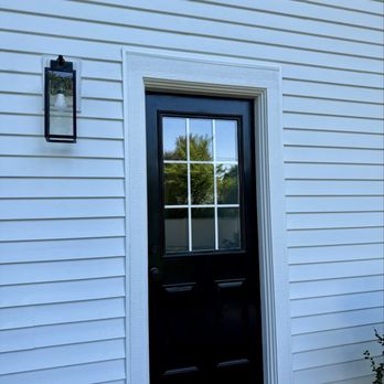 Advanced Windows & Siding - The image shows a black exterior door with a window divided into four panes, set within a white house with horizontal siding. A black outdoor wall-mounted light fixture is located to the left of the door. The house's siding is evenly spaced, and the door frame is white, contrasting with the black door. The reflection in the window shows trees and a blue sky, indicating an outdoor setting. - Windows Installation Near Me - Batavia, Illinois