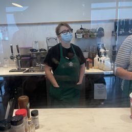 The manager at the Starbucks - A woman with glasses and a face mask stands behind a Starbucks counter, wearing a green apron, with coffee-making equipment and supplies visible.