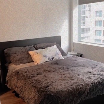 Spotlesstaff - A bedroom features a bed with a plush, dark gray blanket and pillows, positioned near a large window showing a cityscape with tall buildings. - Home Cleaning Near Me