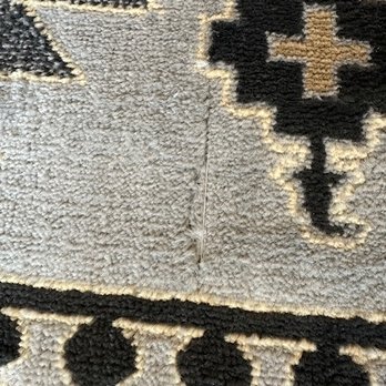 Joybird - A close-up image of a rug featuring geometric patterns in black, beige, and gray. The rug has a visible tear or fold running vertically through the center of the image, affecting the appearance of the design. The texture of the rug's fibers is apparent, highlighting its woven construction. - Rugs Near Me - Denver, Colorado