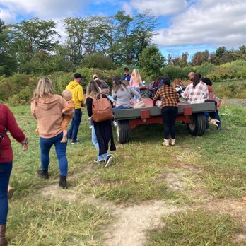 Apple Dave’s Orchards - Multiple individuals, including adults and children, are assembled around a flatbed trailer situated on a grassy area near a pond. The scene features a mix of casual clothing, backpacks, and some people leaning or sitting on the trailer. The background includes trees, bushes, and a partly cloudy sky, indicating an outdoor recreational or educational activity. - Fruits & Veggies Near Me - Warwick, New York