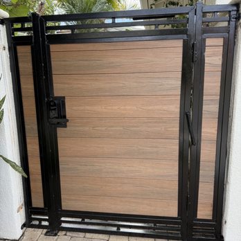 Socal Gates - A black metal driveway gate with wood panel inserts is installed outdoors, with a brick pathway and greenery in the background. - Fences & Gates Near Me - Orange, California
