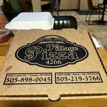 Village Pizza - Our left over pizza - A cardboard pizza box from Village Pizza is positioned on a wooden table inside a restaurant. The box displays the restaurant's name, logo, and contact information. In the background, there are customers seated at tables, including a person wearing a hat and a person in a green shirt, with a counter and kitchen area visible. The setting suggests a casual dining environment with indoor seating. - Pizza Near Me - Corrales, New Mexico