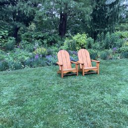 Garden - Two wooden Adirondack chairs are positioned on a green lawn in front of a garden filled with flowering plants, shrubs, and trees. The garden features purple, white, and green foliage, with taller trees in the background providing shade. The scene is tranquil and natural, with the chairs inviting relaxation amidst the garden setting.
