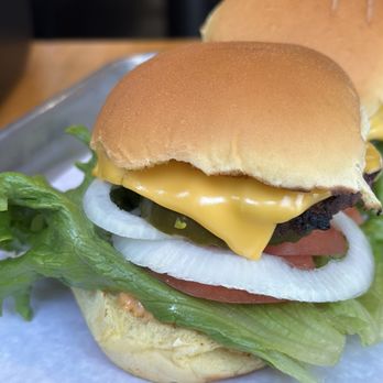 BurgerIM - Sliders - A cheeseburger features a beef patty topped with melted cheese, fresh lettuce, tomato slices, onion rings, and pickles, all inside a soft, toasted bun. The burger is presented on a metal tray with a piece of parchment paper underneath, with another bun visible in the background. - Burgers Near Me - Brambleton, Virginia