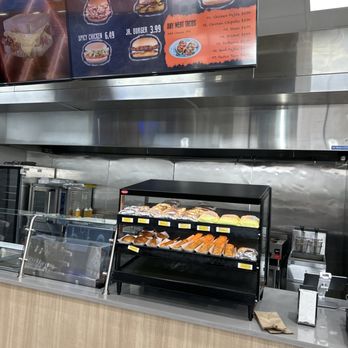Valero - a counter with a variety of food items - A black, three-tier display case on a counter contains sandwiches wrapped in paper, with some unwrapped showing bread and fillings. Behind the counter is a stainless steel kitchen area with appliances and supplies. Above, a digital menu board lists burger and taco options, including cheese burger, hamburger, spicy chicken, Jr. burger, and various tacos, with prices and additional toppings. The setting appears to be a fast-food restaurant or food court. - Convenience Stores Near Me - Pflugerville, Texas