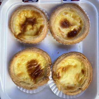 Phu's Bakery - Four Portuguese egg tarts with flaky, golden-brown crusts and creamy custard filling topped with caramelized sugar are arranged in a foam container. The tarts are placed on white paper liners, and the container is set on a surface with a geometric pattern in red, white, and blue colors. - Bakeries Near Me - Rosemead, California