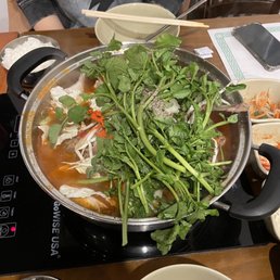 A large stainless steel hot pot containing broth, leafy greens, bean sprouts, and other vegetables is positioned on an induction stove on a wooden table. Surrounding the hot pot are bowls of rice, side dishes, and utensils, with a person seated nearby wearing a dark hoodie with a lightning bolt design. The setting appears to be a Korean restaurant with traditional dining elements.