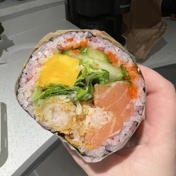 Subo Sushi Burritos - Carribean Sushi Burrito - A sushi burrito with rice, fish, cucumber, lettuce, and toppings is held in a person's hand, showing its cross-section. - Sushi Bars Near Me - Eugene, Oregon