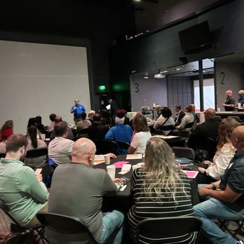 Golfinity - Attendees are seated at tables in a dark auditorium, listening to a speaker at the front, with some using electronic devices. - Golf Lessons Near Me - Austin, Texas
