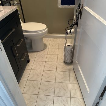 López Construction - Before with white tiles and replaced with blue tile. - A compact bathroom features a white toilet, a dark-colored cabinet, and a towel rack with a floral-patterned towel. The floor is tiled with beige and white patterned tiles. - Masonry/Concrete Near Me