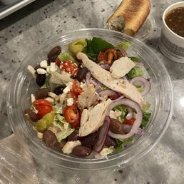 A fresh mixed salad in a clear plastic bowl contains grilled chicken strips, black and green olives, cherry tomatoes, and sliced red onions, placed on a marble countertop. Nearby, there is a piece of bread on a small plastic plate, a cup of soup or broth, and a piece of bread on a small plastic plate. The setting appears to be a casual dining or takeout environment.