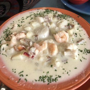 390 Main Street - Seafood Chowder - A ceramic bowl filled with seafood chowder featuring shrimp, potatoes, and herbs in a creamy broth. The dish is garnished with chopped herbs and is placed on a plate. A small cup of tea or broth is visible in the background, along with a blue saucer. The setting appears to be a restaurant table. - Seafood Near Me - Gorham, Maine