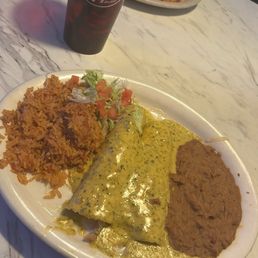 Chicka-chicka Boom-boom Enchiladas - A white oval plate holds Mexican food including seasoned rice, a green sauce-covered enchilada, and a serving of refried beans. A small portion of salad with lettuce and tomato is also visible. In the background, a tall red Dr. Pepper glass with a straw is on a marble table, with another plate of similar food partially visible behind it.