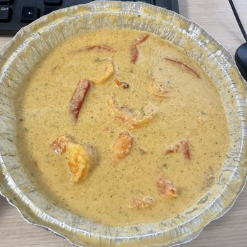 Mangú - Camarones en salsa de coco - A disposable aluminum container contains shrimp in a coconut sauce, with visible pieces of shrimp and red pepper slices. The container is placed on a light-colored desk surface, adjacent to a black computer keyboard and a black mouse. The image appears to be taken in an office or workspace setting. - Dominican Near Me - Boston, Massachusetts