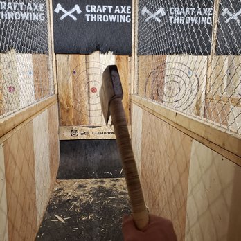 Craft Axe Throwing- Springfield - The image shows a person holding an axe with a wrapped handle, positioned in front of a wooden target with concentric circles and a red bullseye. The setting is an indoor axe throwing lane at Craft Axe Throwing in Springfield, MO, with wooden walls and chain-link fencing on either side. The background features black banners with the Craft Axe Throwing logo and name. The floor has wood shavings and debris from axe throwing activities. - Axe Throwing Near Me - Springfield, Missouri