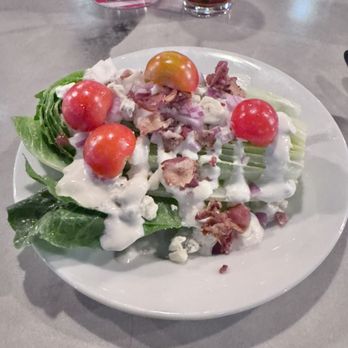 District CR - Half Wedge Salad - A wedge salad featuring iceberg lettuce topped with cherry tomatoes, bacon bits, and a creamy dressing on a white plate. The salad includes chopped onions and crumbled cheese. In the background, two beverages are visible: one in a glass with a straw and another in a glass with a dark-colored drink and a straw. The setting appears to be a restaurant or dining area with a gray table surface. - Bars Near Me - Cedar Rapids, Iowa