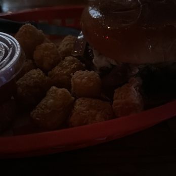 Hideout Pub - Mean green Burger and tots. - A burger with a bun and toppings, served with tater tots and a drink in a dimly lit restaurant setting. - Pubs Near Me - Austin, Texas
