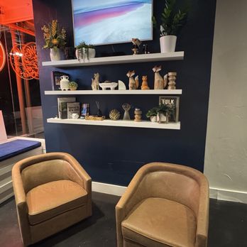 Sans Bar - Two brown armchairs are placed on a dark floor in front of a dark blue wall with three white shelves. The shelves display decorative objects, including vases, figurines, and framed pictures. A flat-screen TV is mounted above the shelves, showing an abstract landscape. To the left, a window with a bench seat is visible, with a neon sign and reflections from outside. The setting appears to be a lounge or waiting area within an indoor space. - Bars Near Me - Austin, Texas