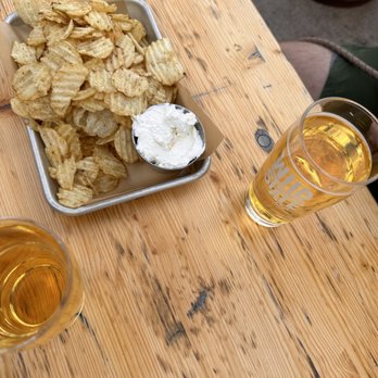 Wild State Cider - Kettle chips + Top the Tater dip - Potato chips served with a creamy dip in a metal tray, accompanied by two glasses of beer on a wooden table. - Cideries Near Me - Duluth, Minnesota