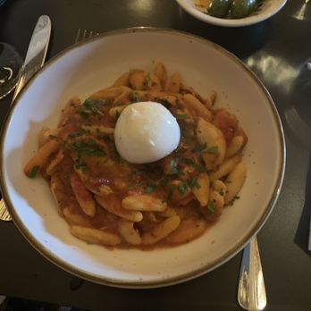 Osa - Cavatelli with roasted Green Circle chicken, spicy tomato sauce and calabro burratina cheese - A large white bowl contains pasta with tomato-based sauce, garnished with chopped herbs and topped with a poached egg. The dish is served on a dark table setting with a fork, knife, and additional small dishes, including a bowl with green olives. The background shows glasses and other tableware, indicating a restaurant dining environment. - Cocktail Bars Near Me - Middletown, Connecticut