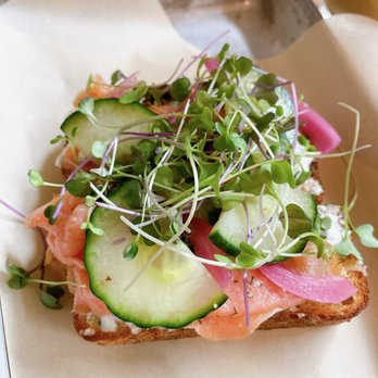 Daycamp Coffee - Lox toast - A piece of toasted bread is topped with smoked salmon, thin cucumber slices, pickled red onions, and a generous handful of microgreens. The toast is placed on parchment paper on a metal tray, with a wooden table surface visible in the background. The presentation emphasizes fresh ingredients and a casual, modern style of serving food. - Coffee Roasteries Near Me - Chico, California