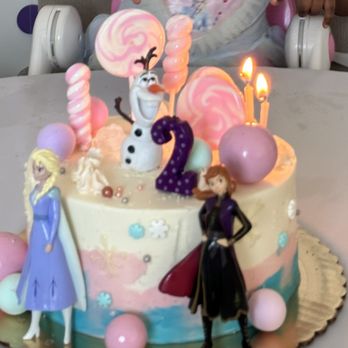 The Evercake - Frozen Theme Cake - A birthday cake decorated with Disney characters Elsa, Anna, and Olaf from Frozen, surrounded by pink and purple candies, lollipops, and a purple number 2 topper. The cake has pastel-colored icing with decorative elements and two lit candles on top. The background shows part of a child's high chair and a person in a light-colored shirt. - Custom Cakes Near Me
