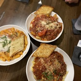 Chicken Parm Sandwich - Three white plates of pasta dishes are on a dark wooden table, each topped with sauce and grated cheese, accompanied by slices of garlic bread. A glass of water is placed on a coaster, and a person’s hands are visible in the background. The setting appears to be a restaurant with a casual dining atmosphere.