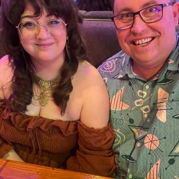 Jazzhaus - a man and a woman sitting at a table - A man and woman are seated at a wooden table in an indoor venue, possibly a bar or restaurant, with other patrons visible behind them. The woman has long, dark, wavy hair, glasses, and is wearing a brown off-shoulder top with jewelry. The man has short, curly hair, glasses, and is wearing a patterned shirt. A smartphone and a pen are on the table in front of them. The background shows a brick wall and a crowd of people, indicating a social or entertainment setting. - Dance Clubs Near Me - Lawrence, Kansas