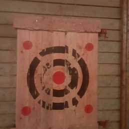 A square wooden target with painted black and red markings is mounted on a wooden wall. The target has a central red circle, surrounded by black concentric circles and additional red dots at the corners. The setting appears to be an indoor axe-throwing venue, with wooden paneling and a rustic atmosphere.