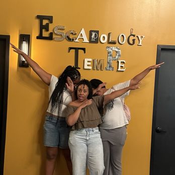 Escapology - Four women stand in front of a yellow wall with decorative letters spelling "Escapeology" and "Tempe." They strike playful poses with arms extended. - Escape Games Near Me - Tempe, Arizona