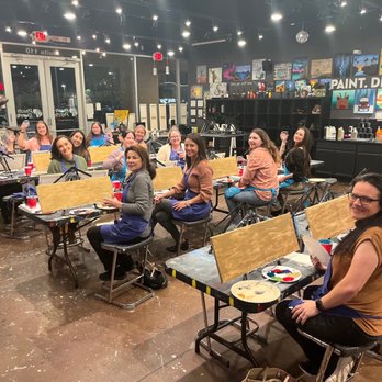 Pinot's Palette - A group of women are seated at tables in a painting studio, participating in a paint and sip event at Pinot's Palette in Gilbert, AZ. The studio has artwork displayed on the walls, and the women are smiling and holding glasses. The space is decorated with string lights and art supplies, with some women wearing aprons. The atmosphere appears social and relaxed. - Paint & Sip Near Me - Gilbert, Arizona