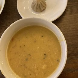 Lentil Soup - A large white bowl contains lentil soup with a yellowish color, visible spices, and a slightly thick consistency. Above the bowl, a white plate holds a steamed dumpling with pleated edges and a small piece of green onion as garnish. The items are placed on a wooden table, suggesting a meal setting at a Himalayan restaurant.